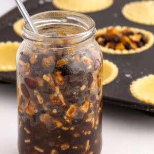 Homemade Christmas mincemeat in a glass jar beside unbaked mince pies in pastry cases.