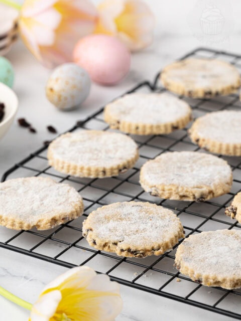 Easter biscuits cooling on a wire rack, dusted with sugar and studded with currants, with pastel Easter eggs and tulips in the background.