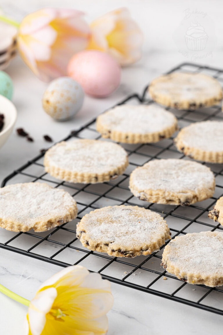 Easter biscuits cooling on a wire rack, dusted with sugar and studded with currants, with pastel Easter eggs and tulips in the background.