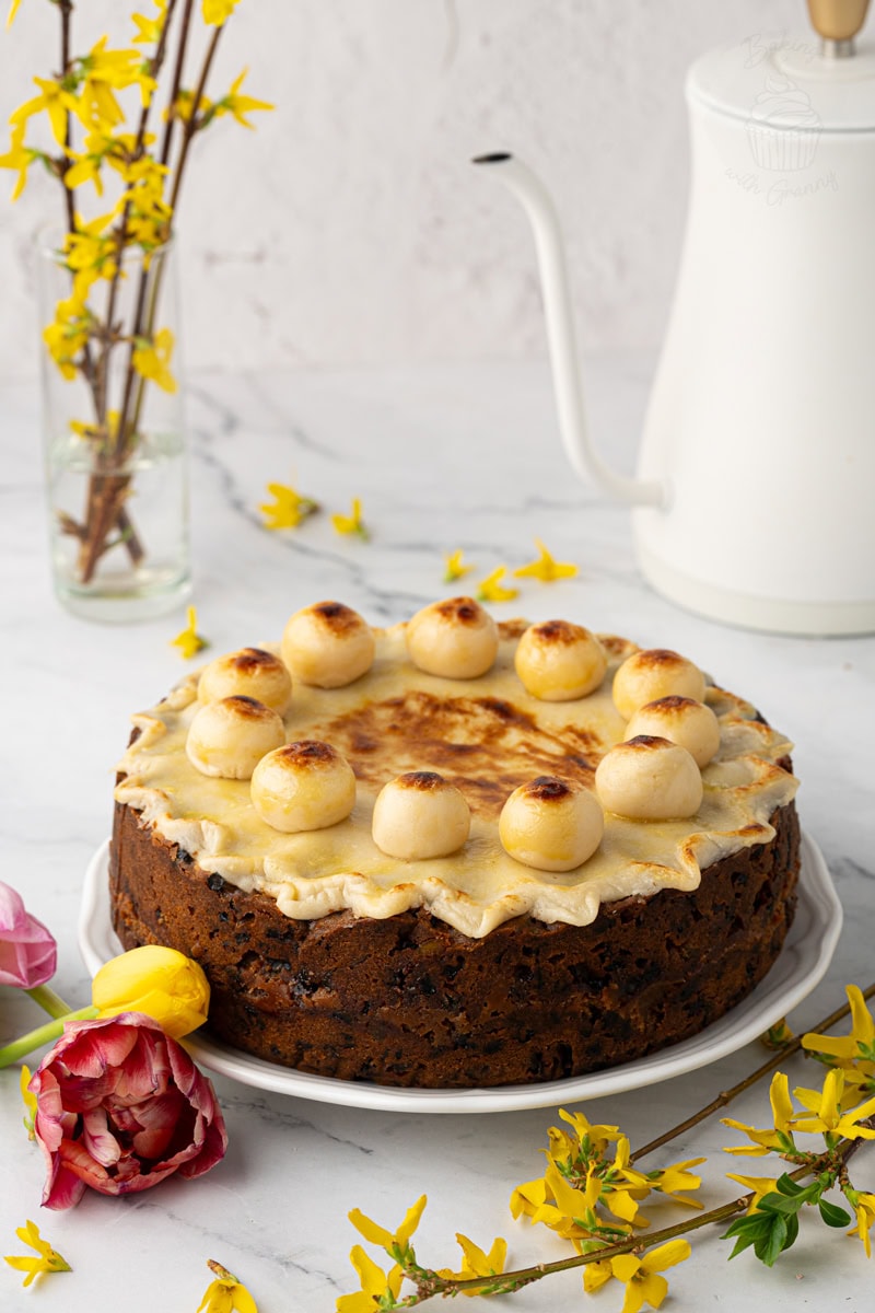 Whole homemade Simnel Cake with marzipan topping and eleven marzipan balls, styled with spring flowers and teapot.