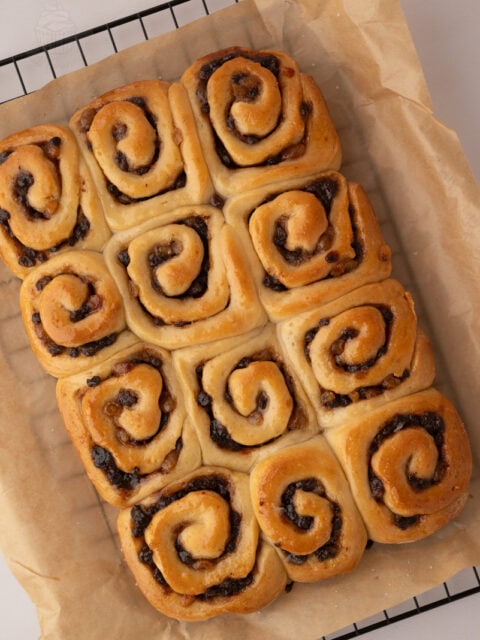 Freshly baked Chelsea Buns cooling on baking paper over a wire rack.