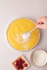 Spoon sprinkling desiccated coconut over homemade Manchester Tart with thick custard filling in a shortcrust pastry case, with glace cherries beside the tart.