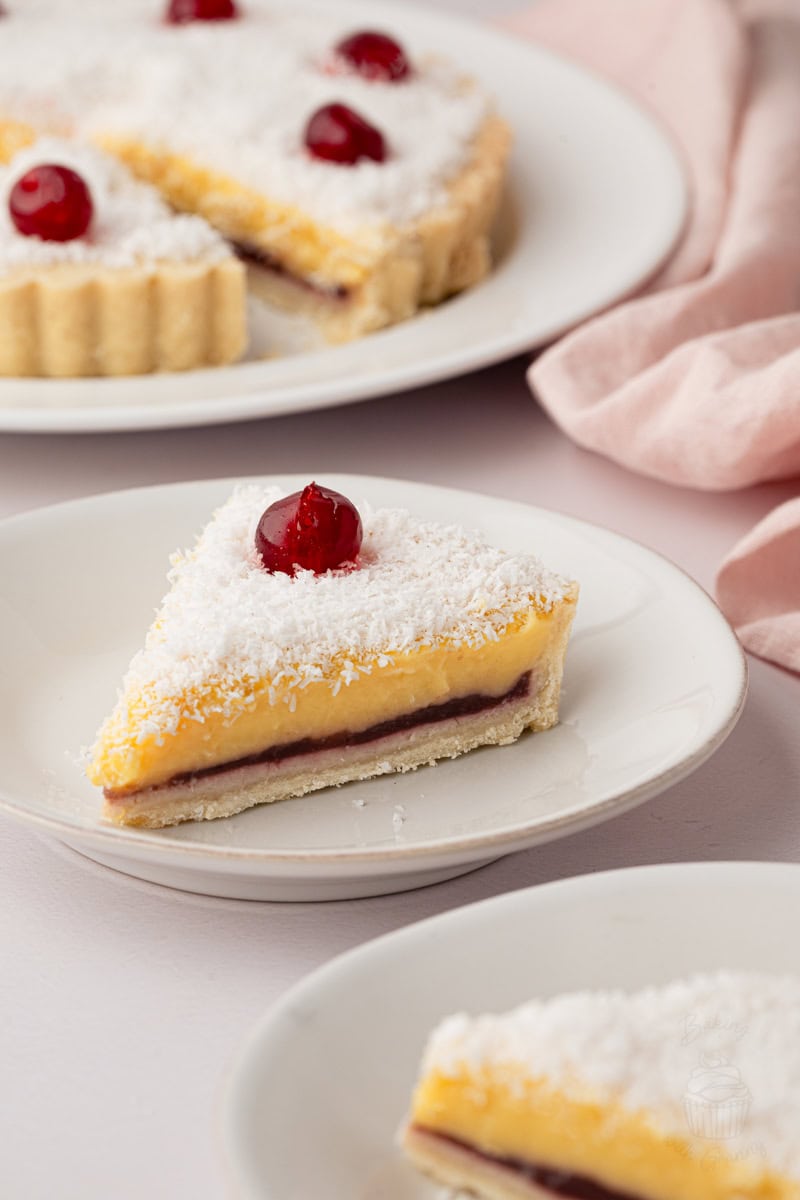 Individual slice of Manchester Tart on a plate with raspberry jam, creamy custard filling, desiccated coconut and a glace cherry topping.