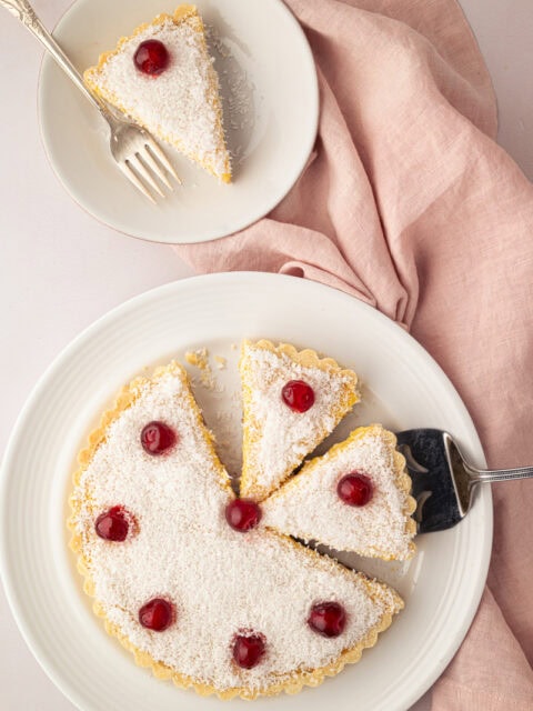 Whole Manchester Tart cut into slices and topped with desiccated coconut and glace cherries on a serving plate.