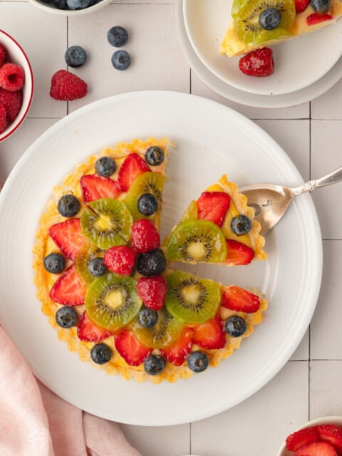 Top-down view of a classic French fruit tart on a white plate, with a silver cake server lifting out a single slice to show the colorful geometric pattern of kiwi, strawberries, and blueberries.