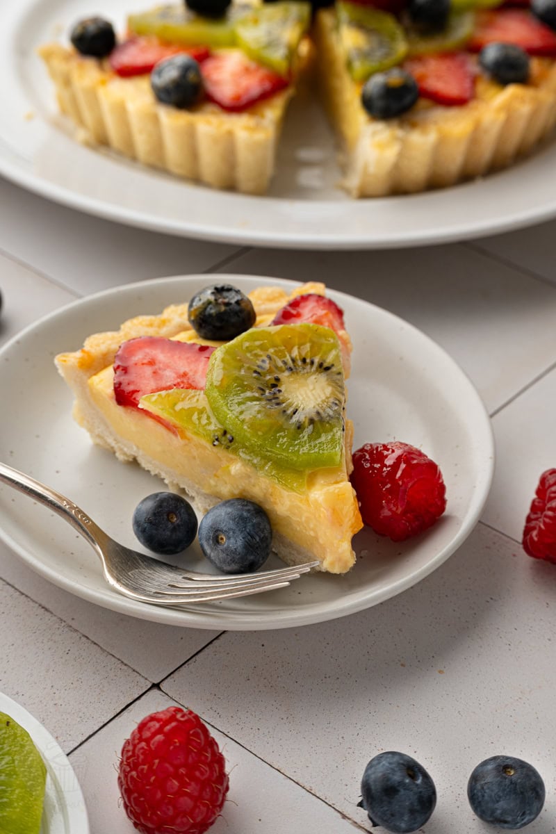 Close-up of a fruit tart slice next to fresh raspberries and blueberries on a white tiled surface, with the remaining whole tart visible in the blurred background.