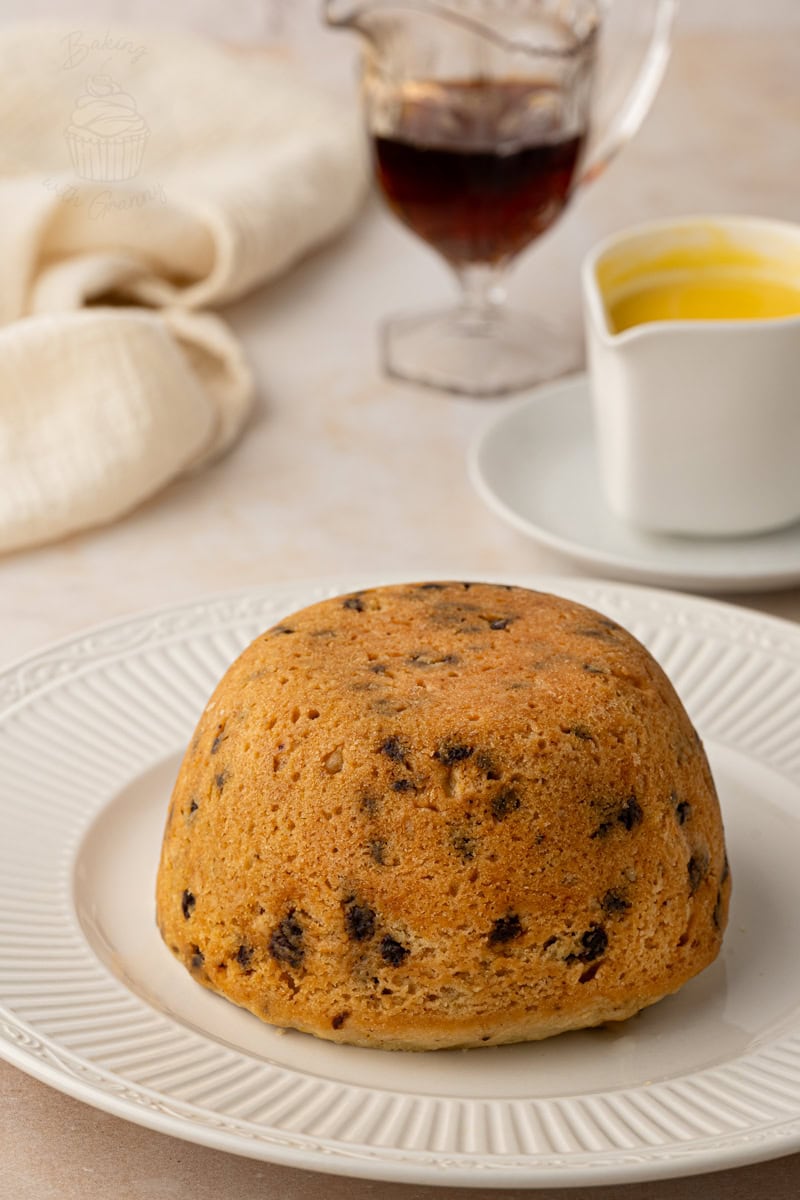 A whole steamed Spotted Dick pudding on a white plate, served with a jug of warm custard and syrup in the background.