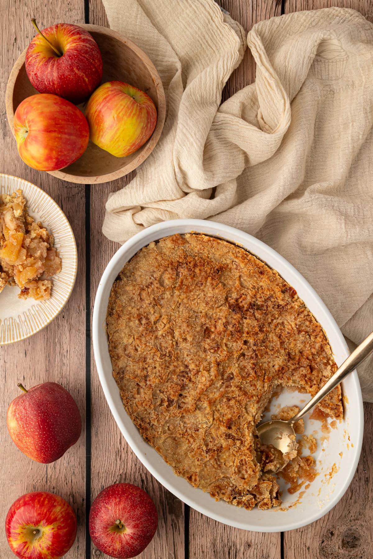 Apple crumble with a portion removed and whole red apples around the baking dish.