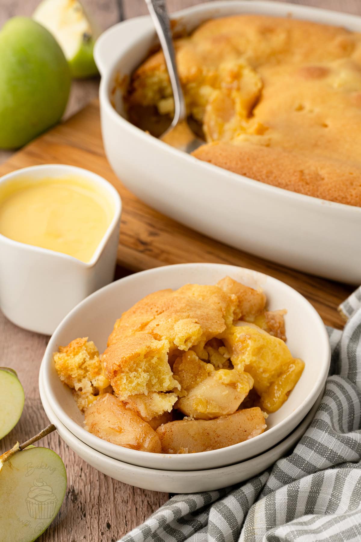 ve’s Pudding with golden sponge in a white baking dish, beside green apples and a jug of custard.