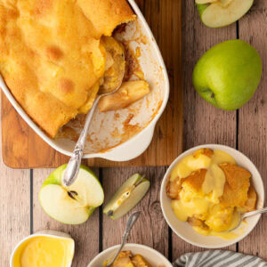 An old-fashioned British Eve's pudding dessert served in a bowl and covered in smooth yellow custard, styled on a rustic wooden background.
