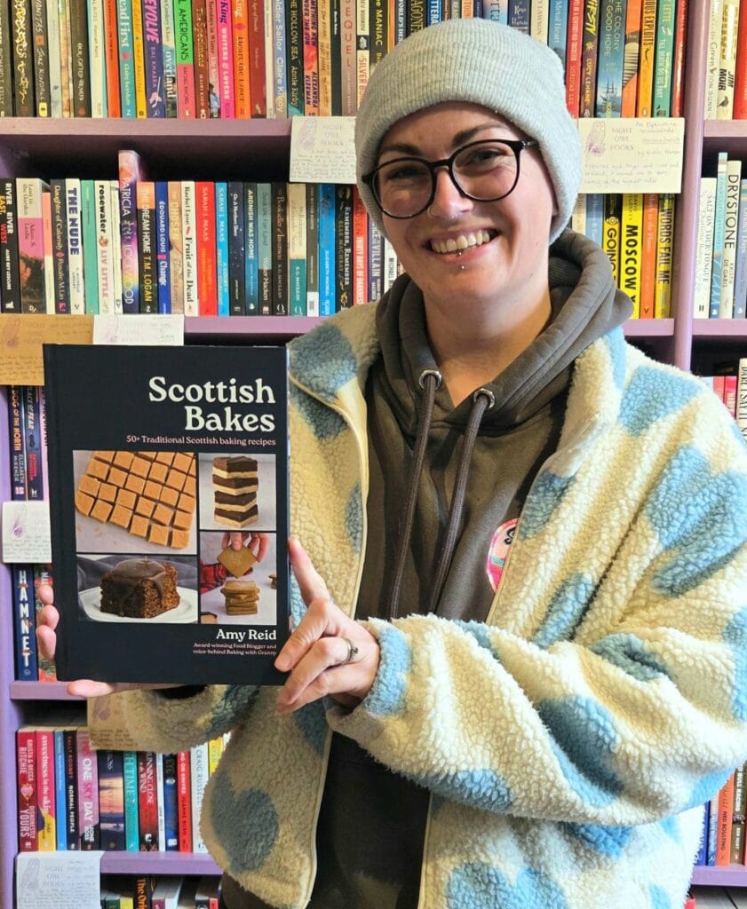 Amy Reid, author of Scottish bakes, holding a copy of her debut cookbook against a book shop backdrop.