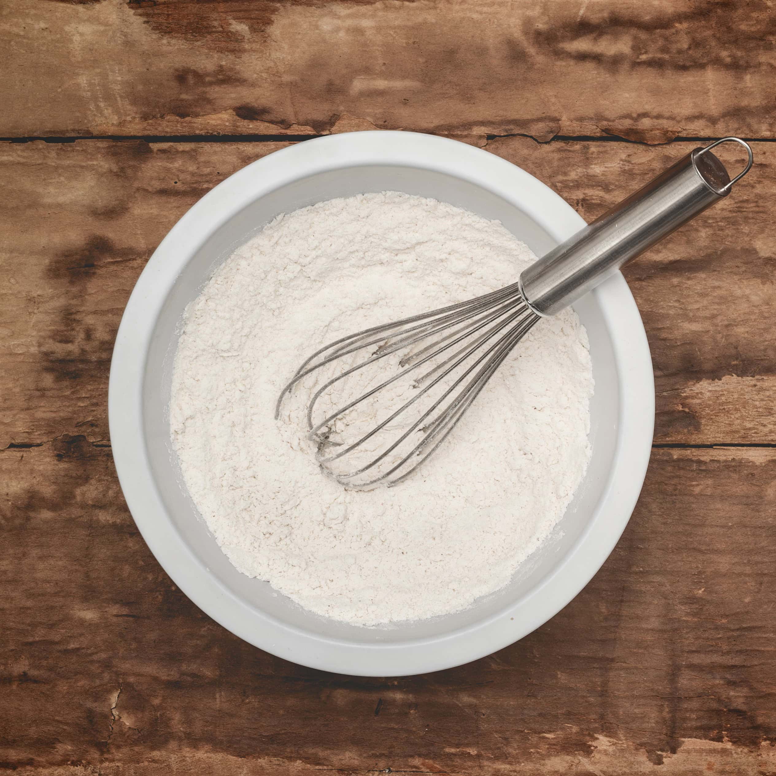 Plain flour (all-purpose flour) in a white bowl with a whisk, on top of a brown wooden surface.