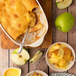 Eve’s Pudding with golden sponge and apple filling, served with custard beside sliced green apples.
