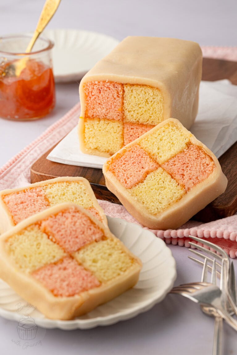 Whole Battenberg cake and sliced pieces showing pink and yellow checkerboard sponge wrapped in marzipan, with apricot jam beside it.