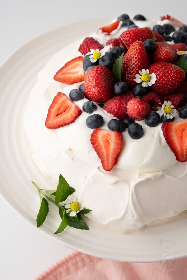 Pavlova topped with whipped cream, strawberries, raspberries, blueberries and small white edible flowers on a plate.