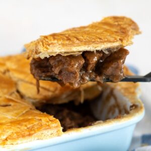 Scottish steak pie with a golden puff pastry topping, with a slice being lifted out.