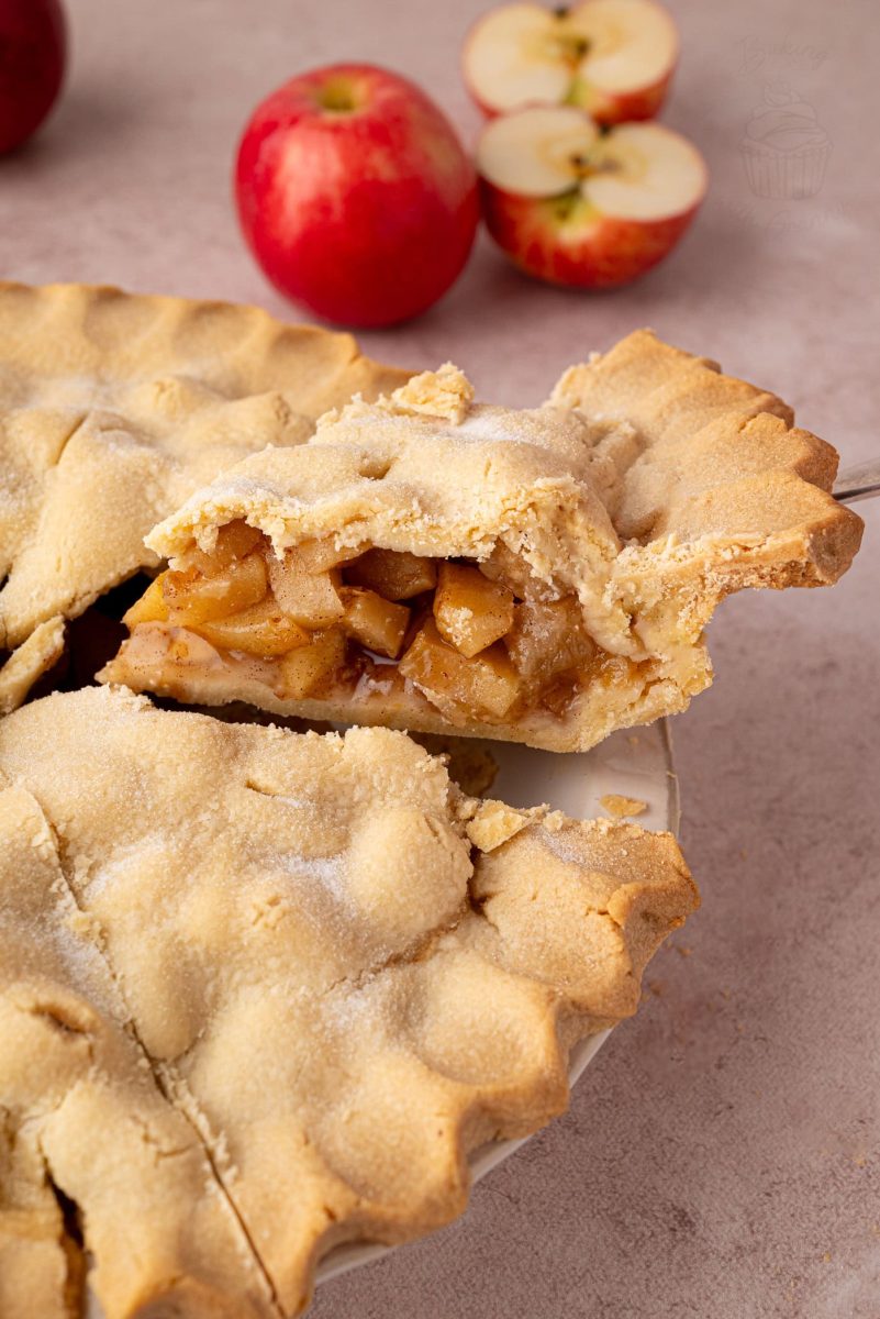 Slice of apple pie on a serving utensil, with whole and halved apples in the background. A traditional apple pie UK recipe ideal for autumn.