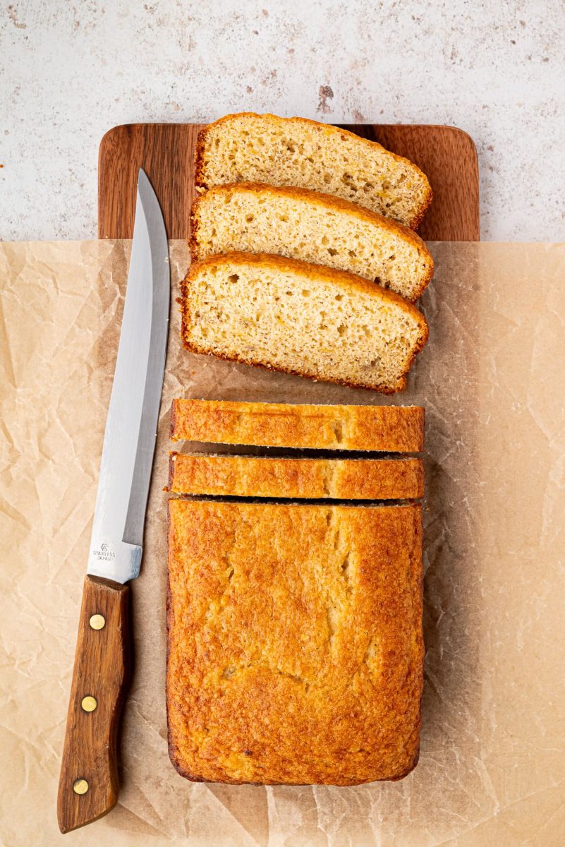 A loaf of sliced banana bread rests on a wooden cutting board, with three slices arranged neatly. A knife with a wooden handle sits on the left. This inviting scene hints at a delicious banana bread recipe against the textured light background.