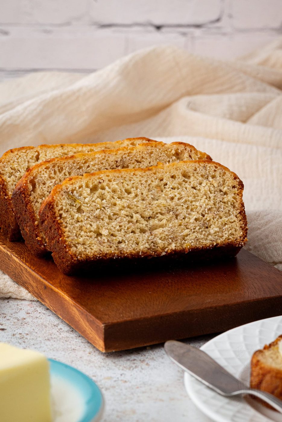 Close-up of sliced banana loaf on a wooden board. The banana bread boasts a golden-brown crust and a moist, textured interior. A plate with a slab of butter and a knife is partially visible in the foreground, complemented by a cream fabric in the background.
