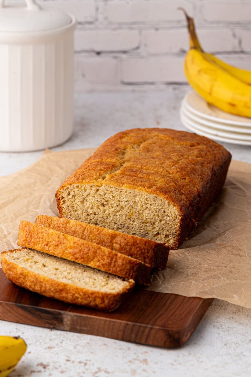 A loaf of banana loaf cake with three slices cut and placed on a wooden board. Ripe bananas, a white canister, and a stack of plates are in the background, all set against a light-coloured brick wall.