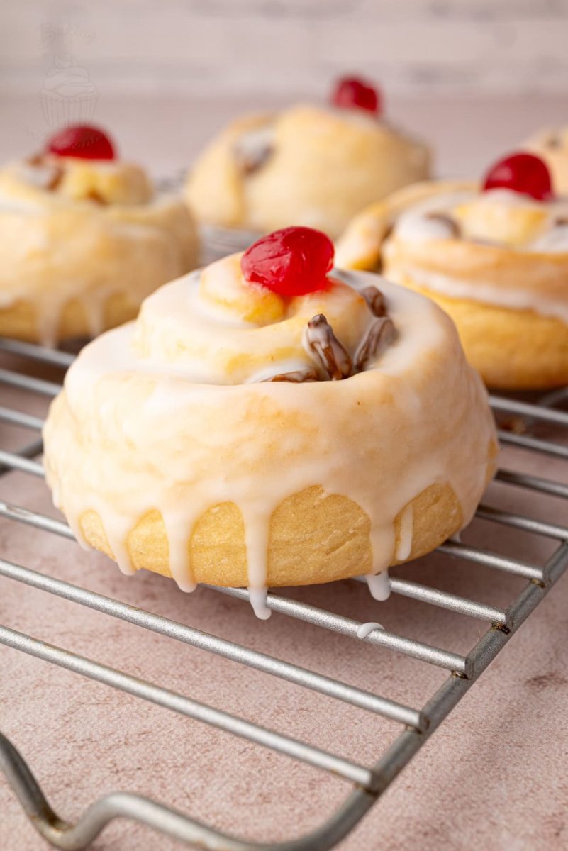 Iced Belgian buns with glacé cherries cooling on a rack, showing the golden bake and soft texture of the buns.