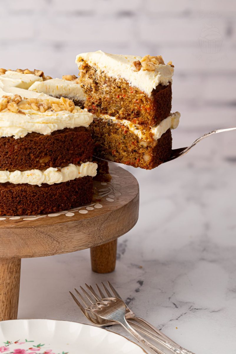 A slice of carrot cake with walnuts and frosting being lifted from a whole cake on a wooden stand, ready to serve.