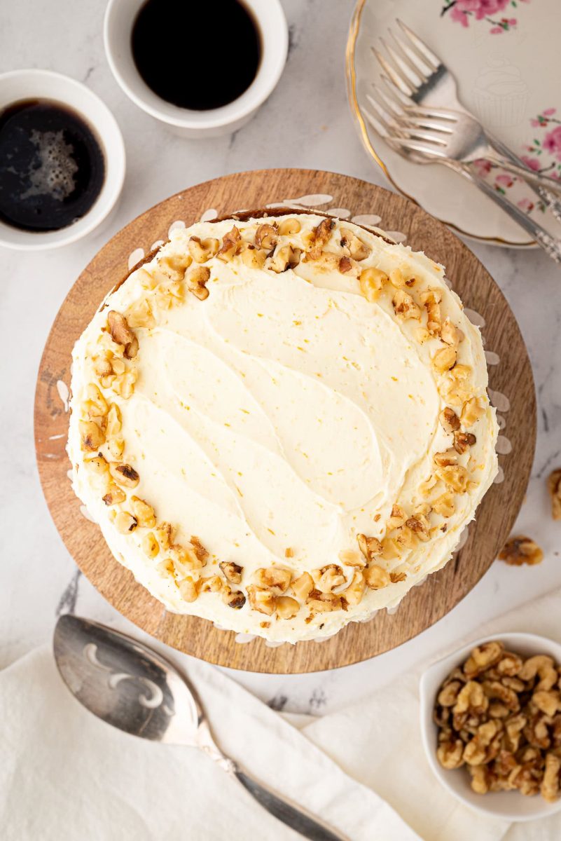 Overhead view of a whole carrot cake with buttercream frosting and chopped walnut topping, surrounded by coffee cups, floral plates, and forks.