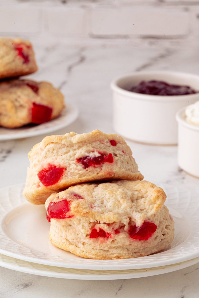A duo of cherry scones sits enticingly atop a white plate, bursting with juicy red cherries. In the background, additional cherry scones are accompanied by small bowls of jam and cream on a sleek marble surface.