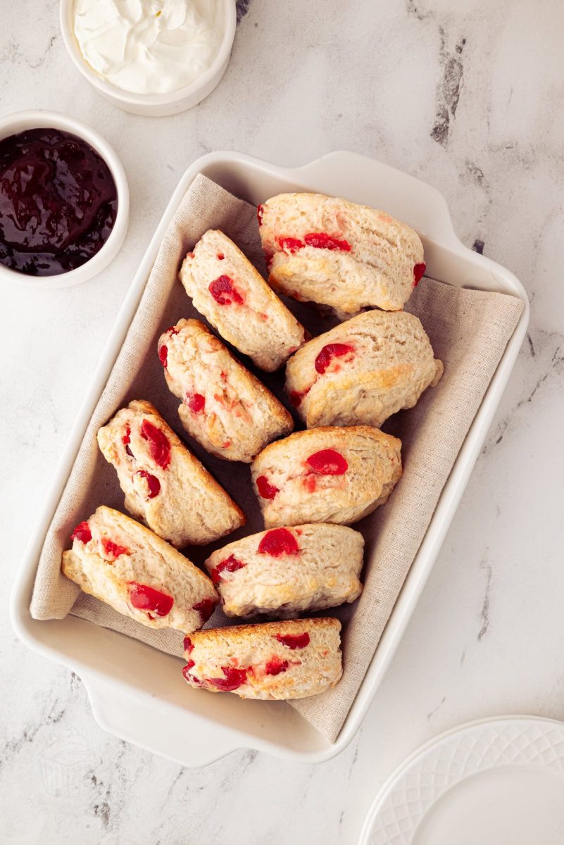 A rectangular dish holds several easy cherry scones with vibrant red glace cherries pieces. The dish is lined with parchment paper. Nearby, small bowls are filled with cream and dark red jam, all elegantly arranged on a light-coloured marble surface.