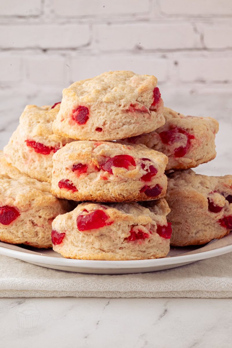 A plate of freshly baked cherry scones featuring bits of bright red glace cherries, stacked in a pyramid shape. The scones have a golden-brown crust and are set against a white brick background, offering an irresistible cherry scones recipe visual delight.