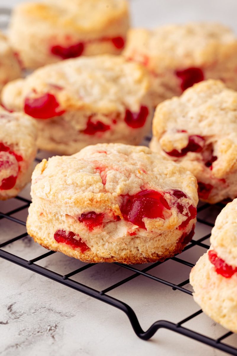 Close-up of cherry scones with visible chunks of glace cherries, placed on a black wire cooling rack. The scones have a golden, crumbly texture, and the cherries add a pop of red colour. The background is a light, textured surface.