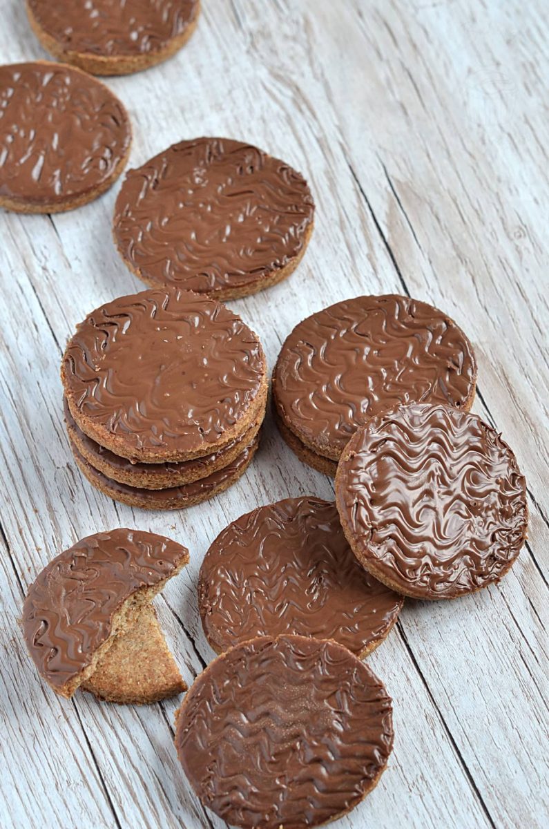 A row of homemade chocolate digestive biscuits on a rustic wooden surface, with one cookie partially eaten to show the crisp, crumbly texture.