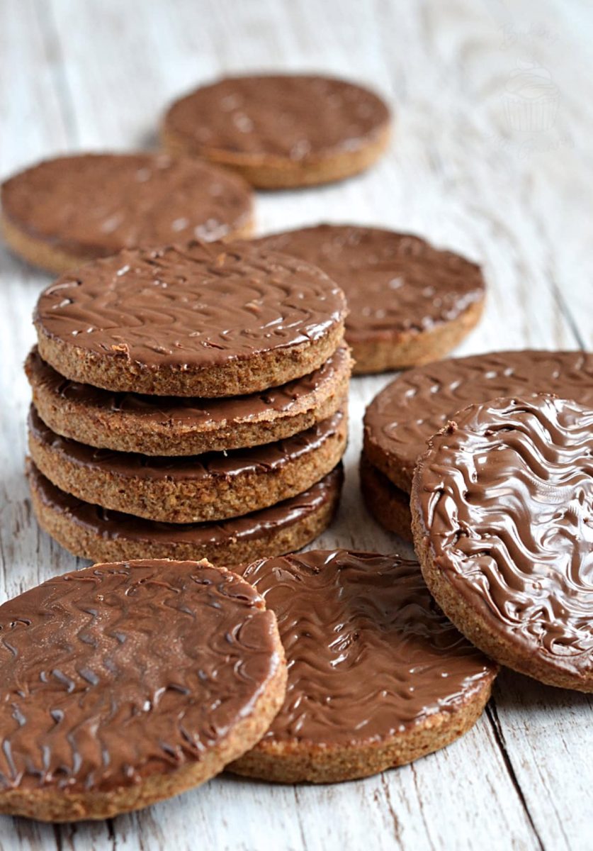 Stacked chocolate digestive cookies surrounded by more on a whitewashed wooden table, showing their glossy chocolate tops and golden brown biscuit base.