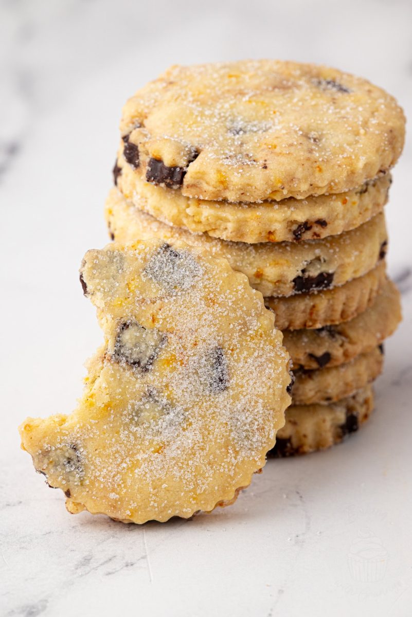 Side view of stacked chocolate orange shortbread biscuits, with a bitten biscuit propped in front to show its crumbly texture and chocolate chunks.