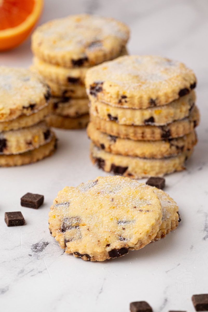 Focus on a single sugared chocolate orange shortbread biscuit, with several stacks of biscuits and an orange half in soft focus behind.
