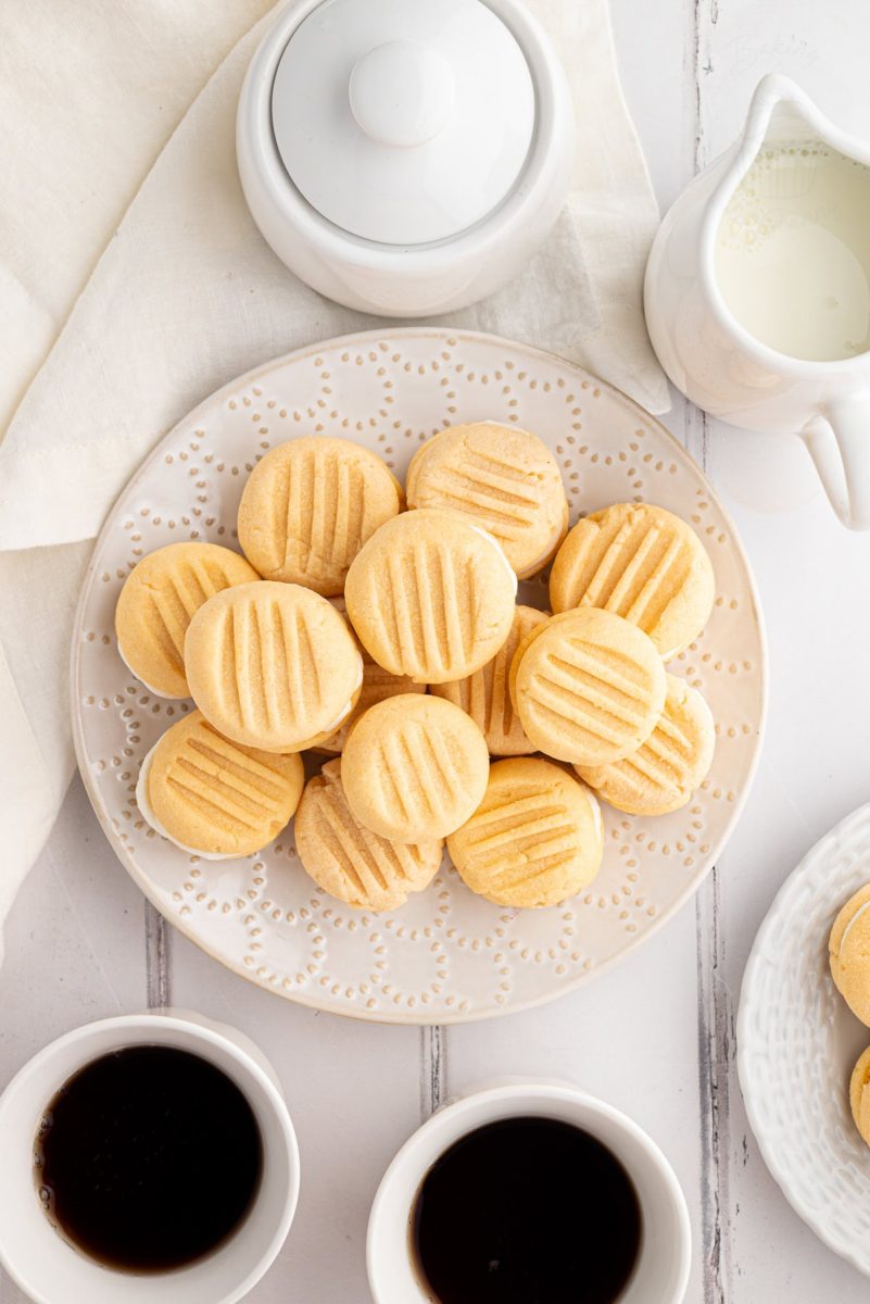 Top-down view of a full plate of custard creams served with cups of coffee and milk jug.