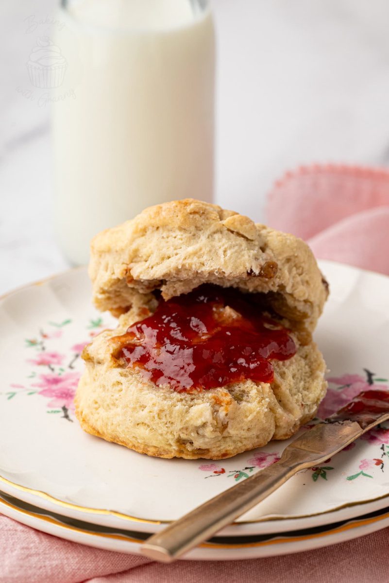 Close-up of a golden fruit scone filled with sultanas, spread with strawberry jam, next to a glass of milk.
