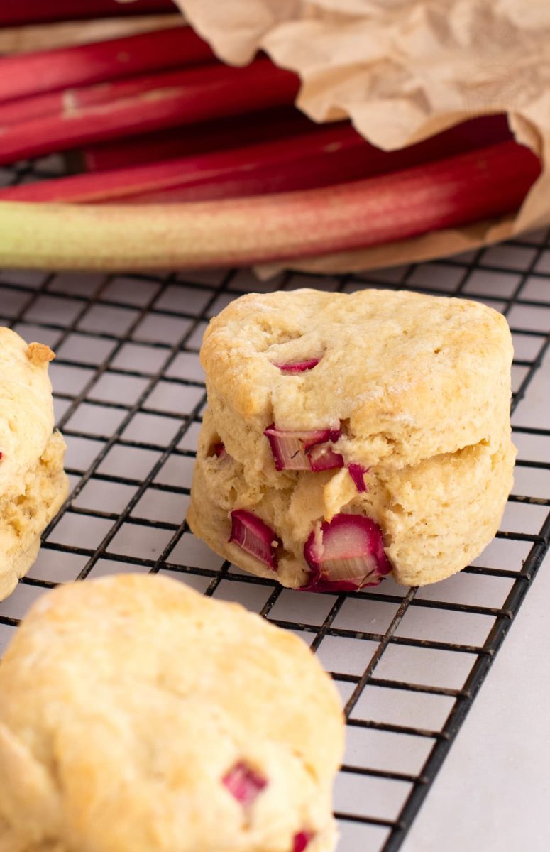 Individual Rhubarb Scone on a cooling rack in front of fresh seasonal rhubarb stems, within a brown paper bag.