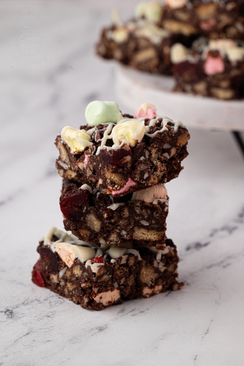Three stacked rocky road squares, complete with visible nuts, marshmallows, and chocolate, are drizzled with white chocolate. They sit on a marble surface like a visual invitation from your favourite rocky road recipe, with more squares blurred in the background.