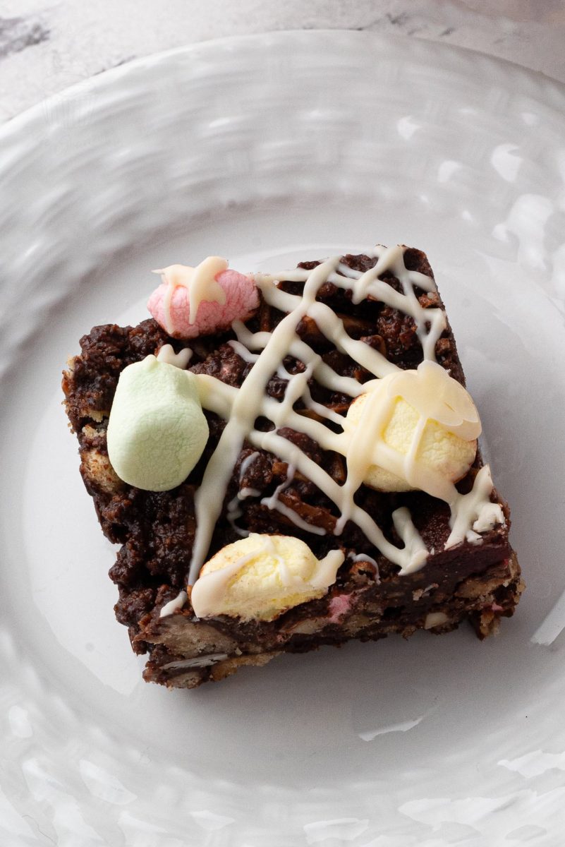 A square slice of rocky road tray bake rests on a white plate, adorned with colourful marshmallows and drizzled with white chocolate in a crisscross pattern.