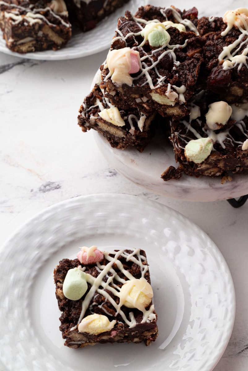 A plate of delightful rocky road squares is topped with colourful marshmallows and a luscious white chocolate drizzle. In the background, another plate offers more of these tempting rocky road treats, all beautifully arranged on a light marble surface.