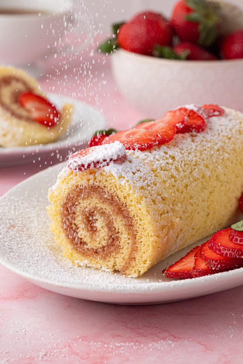 Classic sponge roll cake served on a plate with strawberries and icing sugar, perfect for afternoon tea.