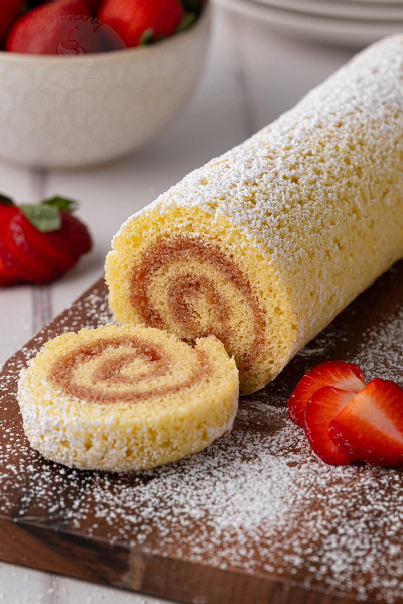 A rolled sponge cake, reminiscent of a classic Swiss Roll recipe, is dusted with icing sugar and sliced on a wooden board. Fresh strawberries are placed around it, with a bowl of strawberries in the background.