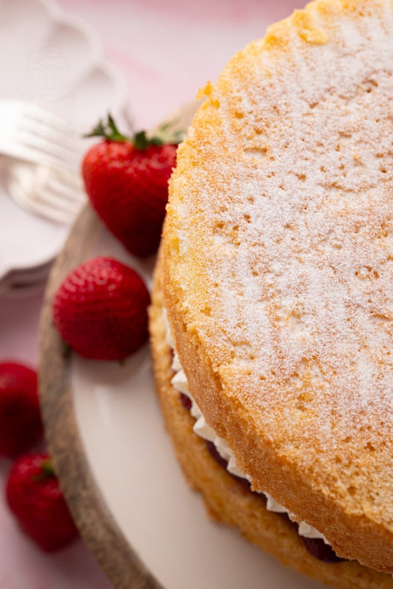 Top-down view of a Victoria Sponge cake dusted with caster sugar, surrounded by strawberries on a rustic cake stand.
