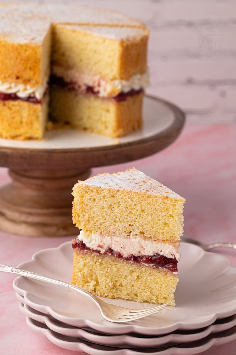 A slice of Victoria Sponge cake on a plate, showing layers of golden sponge, strawberry jam, and buttercream, with the full cake in the background.